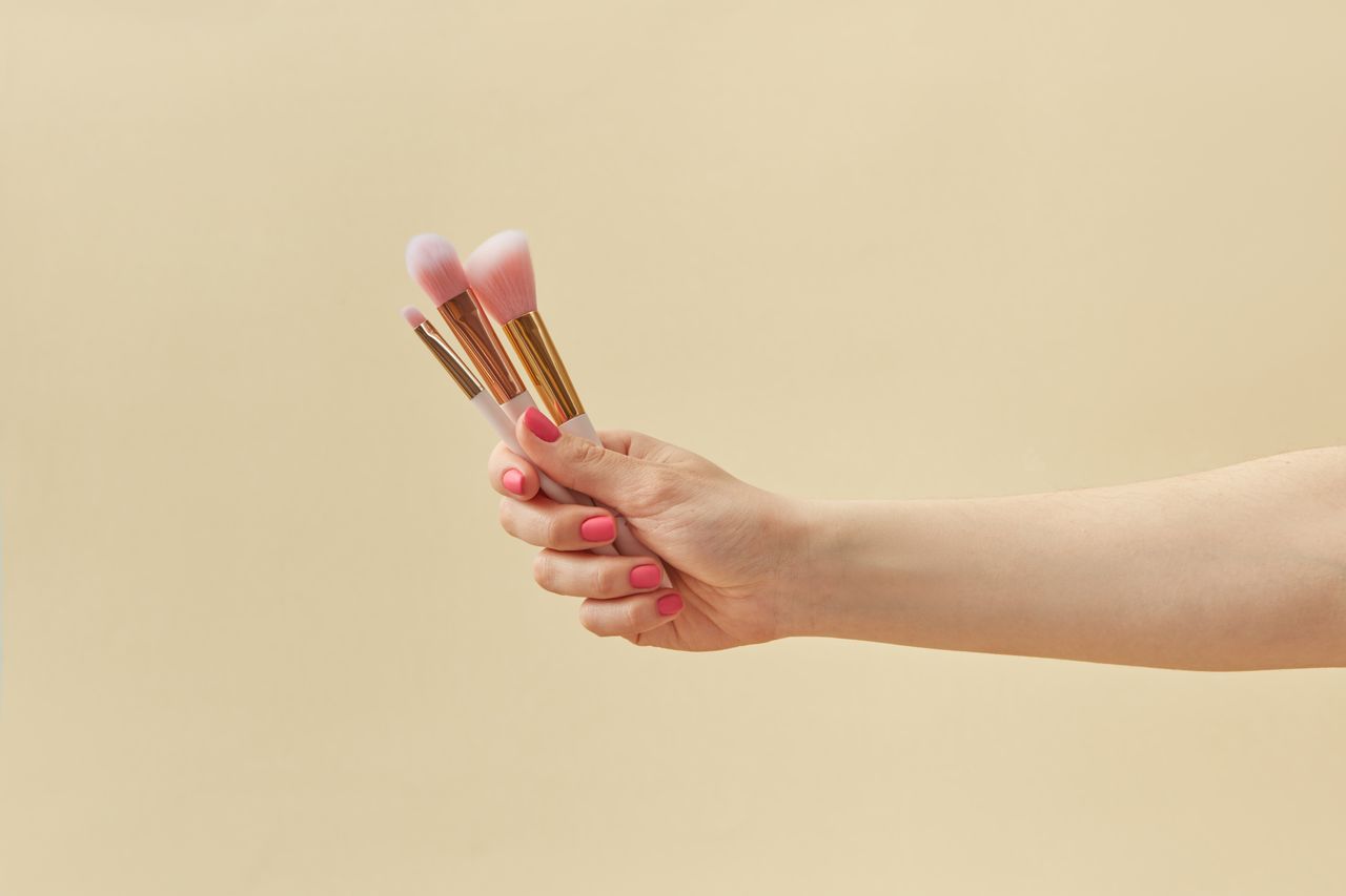 A hand with manicured nails holding makeup brushes against a pink background.
